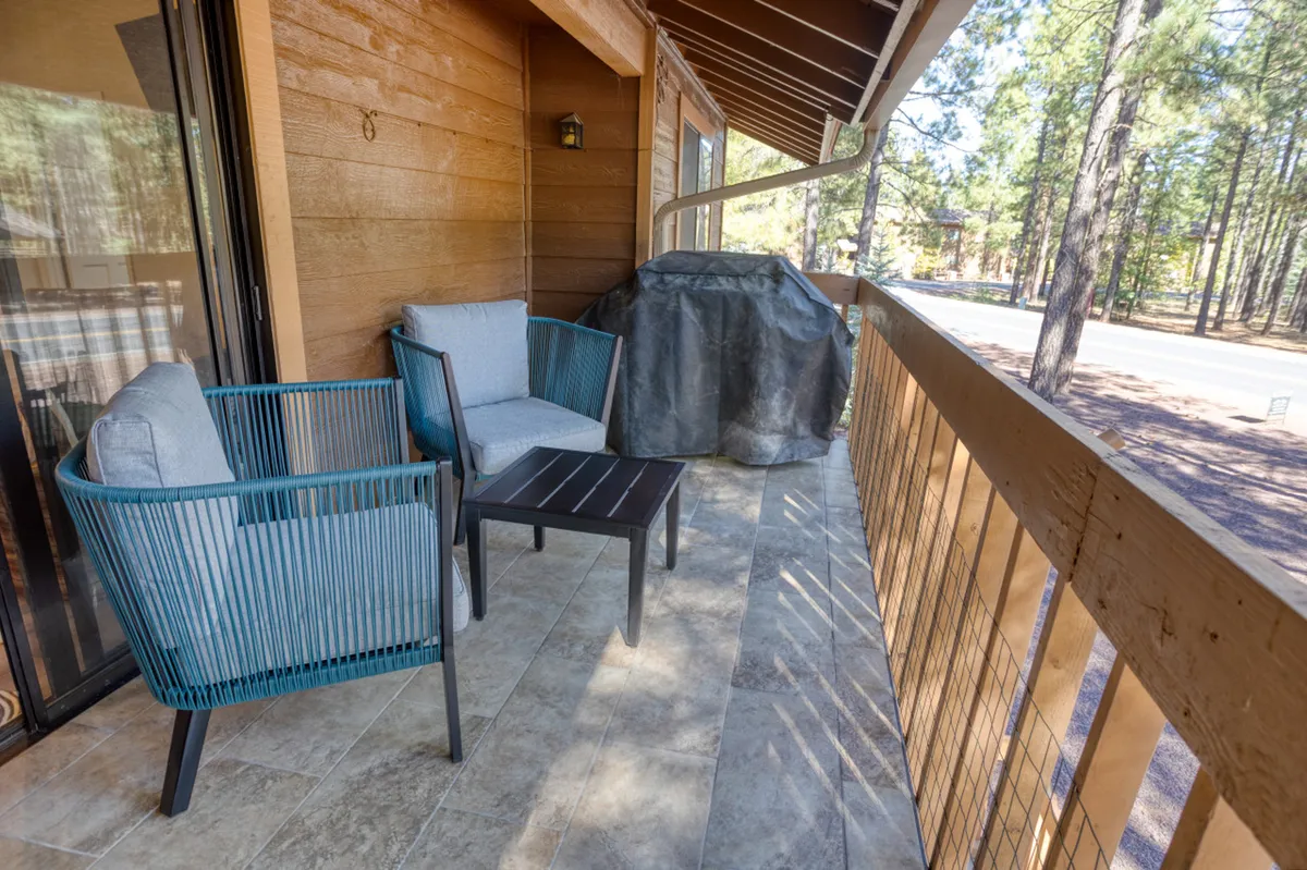 This image shows a spacious covered patio outfitted with two comfortable wicker chairs, a small coffee table, and a barbecue grill. The patio offers tranquil views of wooded surroundings, with natural light illuminating the seating area and grill—perfect for morning coffee, outdoor meals, or relaxed evening gatherings.