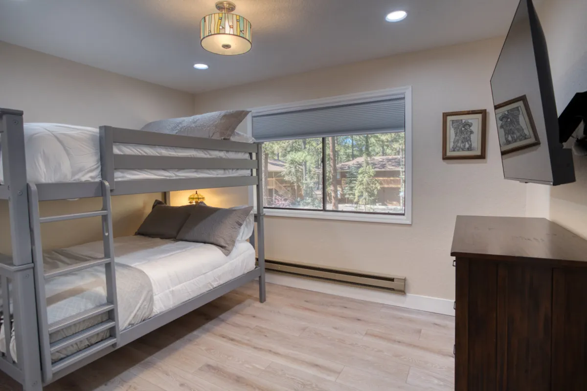 A charming third bedroom featuring a full-over-full bunk bed, a window with sunlight streaming in, a decorative light fixture, and a television. The setup is ideal for children or friends, offering space for sleepovers, relaxation, and shared experiences.