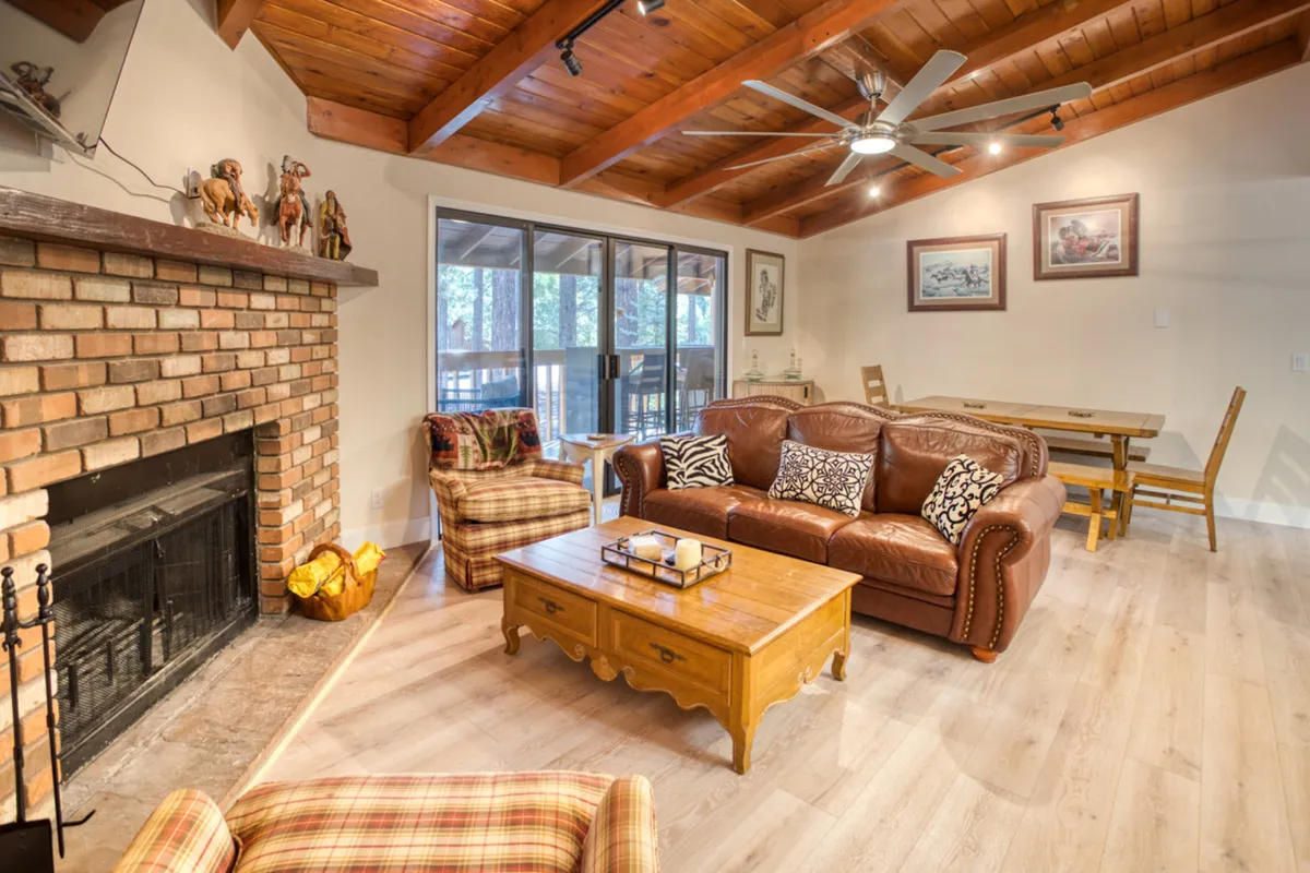 A spacious mountain great room with a brick fireplace as the focal point, flanked by a plush leather sofa and a wooden coffee table. Natural light pours in through large windows, illuminating a warm, welcoming area that seamlessly flows into an adjacent dining space. The overall ambiance is cozy yet open, ideal for gatherings and relaxation.