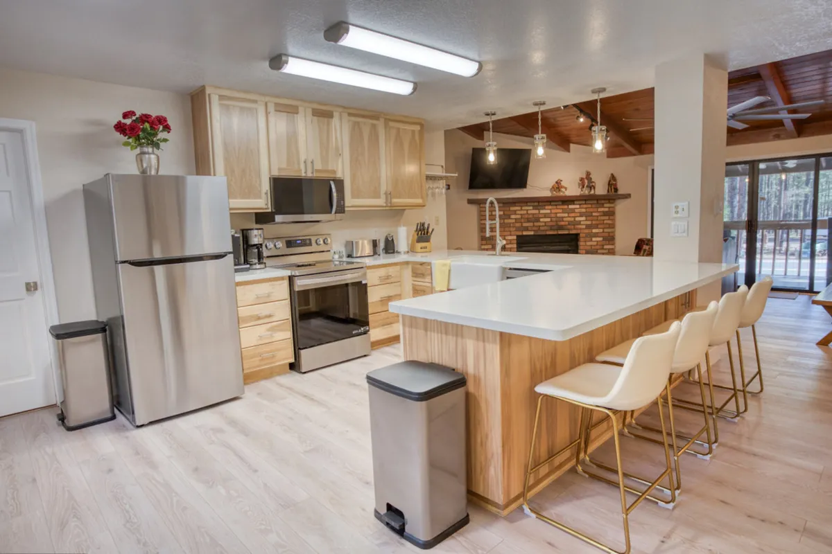 The image showcases the heart of the condo: a spacious, inviting kitchen and living area with a central island, gleaming stainless steel appliances, warm wooden cabinetry, and a cozy brick fireplace. The open layout is perfect for socializing and gathering with family or friends, set in a modern yet welcoming mountain retreat.