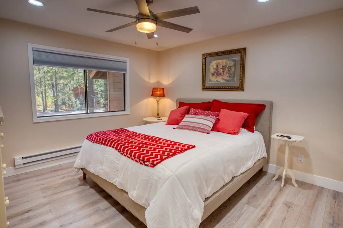 A serene bedroom with red and white bedding, a window offering a tranquil view of the trees, a bedside lamp, and tasteful wall art. The natural light pouring in creates a restful and inviting atmosphere.