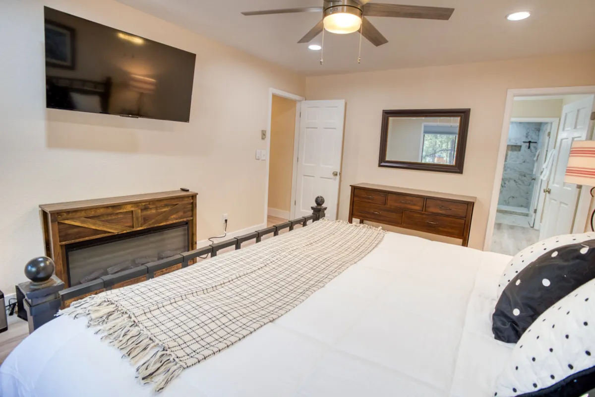 A warmly lit bedroom featuring a comfortable bed adorned with a decorative blanket, a wooden dresser, a wall-mounted TV, a ceiling fan, and a framed mirror. The overall ambiance is inviting and cozy, ideal for relaxation and leisure.